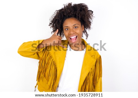 young beautiful African American woman wearing yellow jacket over white wall makes phone gesture, says call me back again, has glad expression.