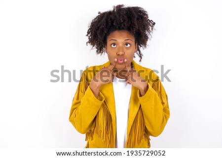 young beautiful African American woman wearing yellow jacket over white wall crosses eyes and makes fish lips funny grimace