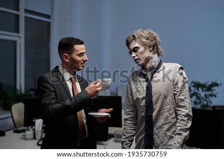 Young elegant businessman offering cup of coffee to zombie colleague in office environment Royalty-Free Stock Photo #1935730759