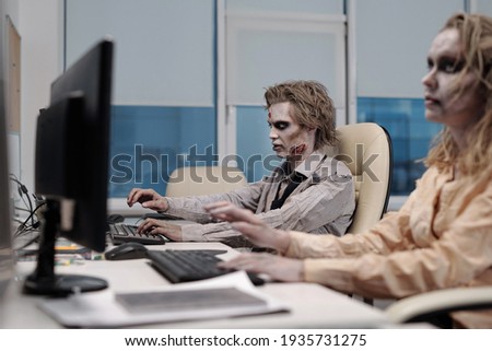 Zombie businessman sitting in front of monitor with dead female colleague working next to him