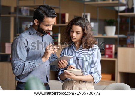 Indian manager holding digital tablet having discussion with latin employee meeting in office lobby. Two diverse professionals talking using pad discussing project strategy, checking corporate plan. Royalty-Free Stock Photo #1935738772