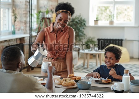 Young happy African woman pouring coffee or tea into mug of her husband by breakfast Royalty-Free Stock Photo #1935744322