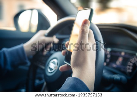 Teen drive a car and use smartphone. Young man reading messages holding a cell phone while driving. Dangerous behavior, accident risk. Danger, transgression, youth, distraction concept. Focus on hand. Royalty-Free Stock Photo #1935757153