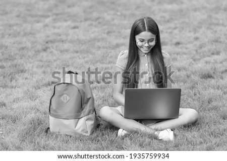 smart happy kid wear glasses using laptop for doing school homework online sitting in park on green grass with backpack, education