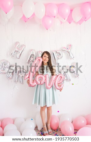 Smiling brunette woman in mint dress holding pink word love. Birthday decorations with white and pink color balloons and confetti for party on a white wall. Happy birthday anniversary. Girls party.