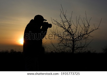 Silhouette of a male photographer with a camera, illuminated by an orange sunset.Unrecognizable. A beautiful landscape with a branching tree.