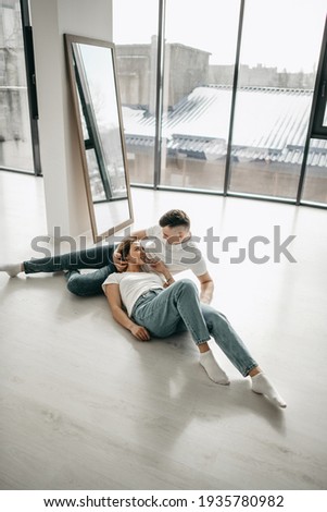 Beautiful young lovers pair of man and women in white t-skirt, blue jeans hugging, laying at home near mirror, windows. Girl blonde, happy people, romance, date, white interior, passion, reflection