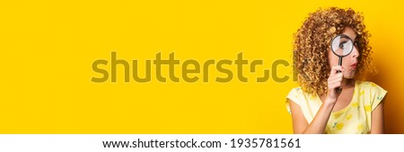 surprised curly young woman looks through a magnifying glass on a yellow background. Banner