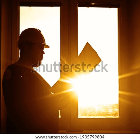 male builder engineer wear security helmet look at blueprint paper construction drawing plan on background of sunset window frame blue yellow sky. window frame.