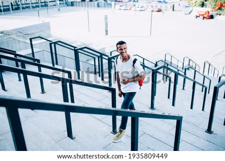 From above of positive young African American man in blue jeans walking upstairs with modern laptop and looking away on sunny day Royalty-Free Stock Photo #1935809449