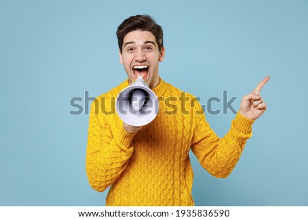 Young student man 20s in casual knitted yellow fashionable sweater scream aside shout in megaphone point index finger aside on copy space workspace area isolated on blue background studio portrait Royalty-Free Stock Photo #1935836590