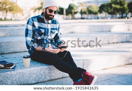 Bearded male blogger dressed in stylish clothing spending leisure time at urban setting listening online podcast, Turkish hipster guy in earphones and sunglasses watching video vlog via touch pad