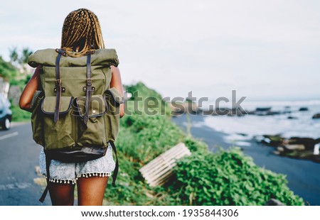 Back view of African American female tourist with backpack exploring nature environment during summer adventure on tropical island, dark skinned active woman with touristic rucksack outdoors Royalty-Free Stock Photo #1935844306