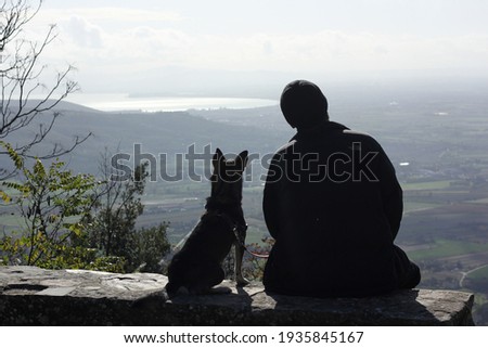 Silhouette of a man and a silhouette of a dog sitting nearby and looking into the distance from the height of the mountain for the beautiful views of Tuscany 