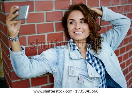 Cheerful hipster girl smiling at camera during leisure at urban setting shooting video vlog for sharing to social media, joyful female blogger posing while photographing herself using mobile app
