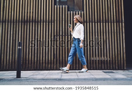 Side view of stylish positive middle aged female in jeans and sneakers strolling along street near modern building and drinking coffee Royalty-Free Stock Photo #1935848515