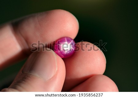 Natural 6 rays star loose red ruby oval cabochon gemstone holded in hand fingers on bright sunny day light. Heated lead glass filled ruby gem. Precious gemstone, corundum Al2O3 Aluminium oxide formula Royalty-Free Stock Photo #1935856237