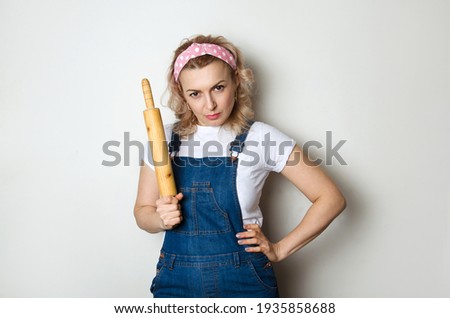A girl with a strict expression, holds a rolling pin in her hands and looks indignantly. For a healthy lifestyle, proper nutrition.Angry woman in the kitchen.Copy space.Serious woman.Dough rolling pin Royalty-Free Stock Photo #1935858688