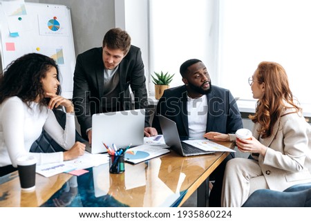 Influential business team in modern office on briefing, working on new project. Successful male and female employees discussing ideas, learning financial graphs, using laptop Royalty-Free Stock Photo #1935860224
