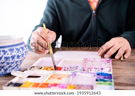 Asian male artist paints with a paint brush and watercolor palette on a laptop face paper.