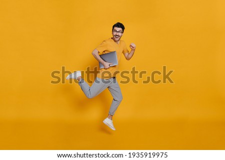 Full length photo of a cheerful funny caucasian smart young man in eyeglasses, stylishly dressed, holding laptop, smiling and jumping high running on isolated orange background Royalty-Free Stock Photo #1935919975