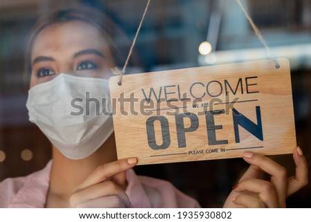 Smiling young asian owner retail,coffee shop woman turning sign board to open wearing face mask,protect to pandemic of coronavirus, reopen store after close lockdown quarantine in covid to new normal.