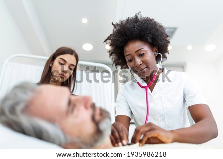 Sick man patient with African American female doctor listens his chest with stethoscope in hospital emergency room. Doctor and nurse come to visit elder patient at hospital room.