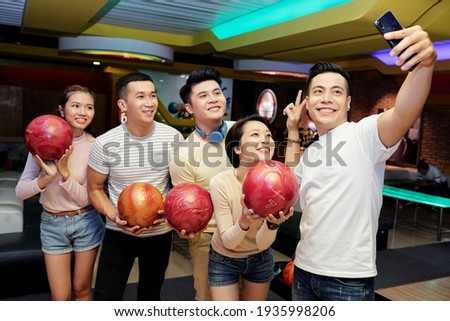 Group of Asian friends posing with balls at camera of mobile phone while playing bowling