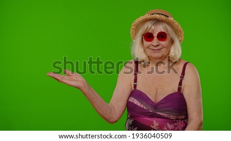 Senior pensioner woman tourist in swimsuit points hand on left at blank space shows advertising content from mobile phone on chroma key background. Old granny traveler model on summer vacations