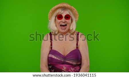 Amazed senior pensioner woman tourist in swimsuit bra, red sunglasses and hat shocked by sudden victory, saying wow on chroma key background. Surprised old granny traveler model on summer vacations