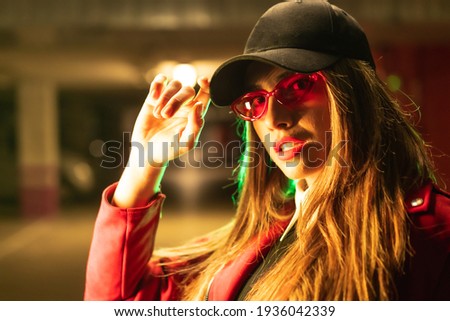Photography with red and green neons in a parking lot. Portrait of a young blond Caucasian woman in a red suit, sunglasses and a black cap