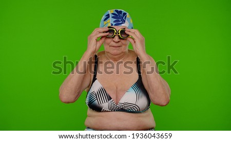 Senior woman tourist wearing swimming glasses, looking at camera, dancing on chroma key background. Travel, summer holiday vacation, beach party. Elderly grandmother in swimsuit, indoor studio shoot