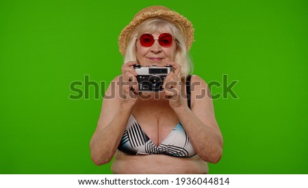 Mature woman photographer traveler in red sunglasses taking picture photos on retro camera. Senior grandmother in swimsuit bra on chroma key background. Tourism, summer holiday vacation, trip to sea