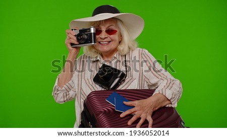 Senior woman tourist photographer taking photos on retro camera and smiling on chroma key background. Travel, summer holiday vacation. Elderly grandmother luggage with luggage indoor studio shoot