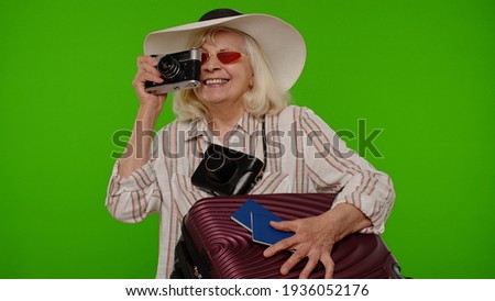 Senior woman tourist photographer taking photos on retro camera and smiling on chroma key background. Travel, summer holiday vacation. Elderly grandmother luggage with luggage indoor studio shot