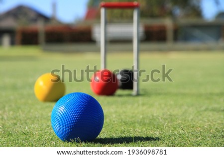 A blue croquet ball is in focus in the foreground of a croquet lawn, while the red, yellow and black balls and the hoop are out of focus in the background Royalty-Free Stock Photo #1936098781