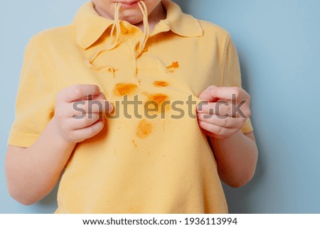 Ketchup stains on a yellow t-shirt. Hungry boy licking snack from his fingers. isolated on blue background. High quality photo Royalty-Free Stock Photo #1936113994