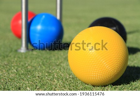 A yellow croquet ball is in focus in the foreground on a grass lawn with the red, black and blue croquet balls and the croquet hoop in soft focus in the background Royalty-Free Stock Photo #1936115476