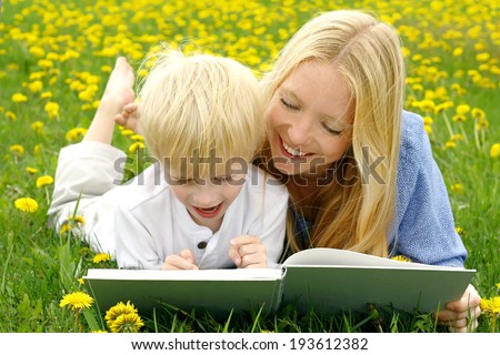 a happy mother is laying in the Dandelion flowers outside with her young child, reading him a story from a book.