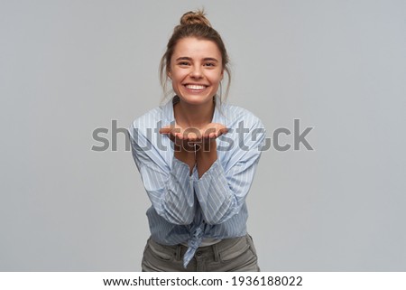 Nice looking woman, beautiful girl with blond hair gathered in bun. Wearing striped knotted shirt. Keeps her palms facing up and broadly smile. Watching at the camera, isolated over grey background