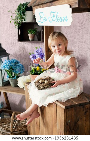 Beautiful blonde girl with nest with eggs on background of decorated flower shop. Easter, flower business concept. Inscription in French "Flowers"