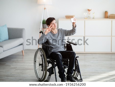 Happy disabled teenager in wheelchair taking selfie on smartphone, smiling at camera indoors