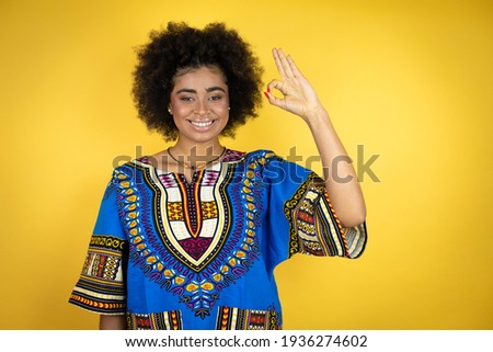 African american woman wearing african clothing over yellow background doing ok sign with fingers and smiling, excellent symbol