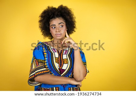 African american woman wearing african clothing over yellow background serious face thinking about question with hand on chin, thoughtful about confusing idea