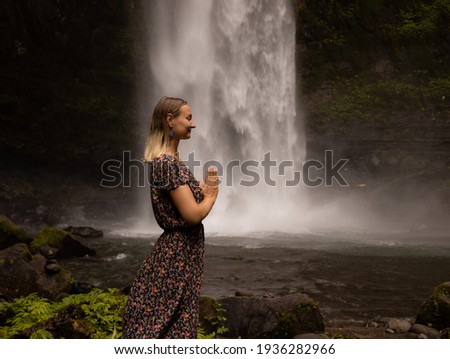 Young Caucasian woman spending time near waterfall. Meditation and relaxation. Hands in namaste mudra. Closed eyes. Travel lifestyle. Woman wearing dress. Nung Nung waterfall in Bali, Indonesia