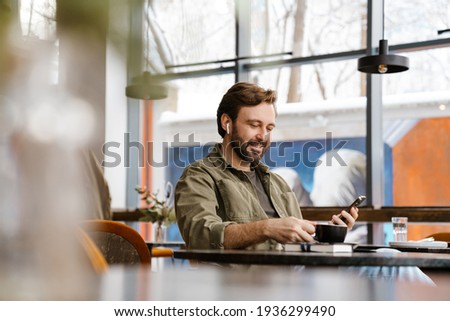 Unshaven happy man in earphones using mobile phone while drinking coffee at cafe Royalty-Free Stock Photo #1936299490