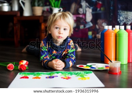 Sweet toddler blond child, boy, painting with colors, little chicks walking around him, making funny prints on his paper