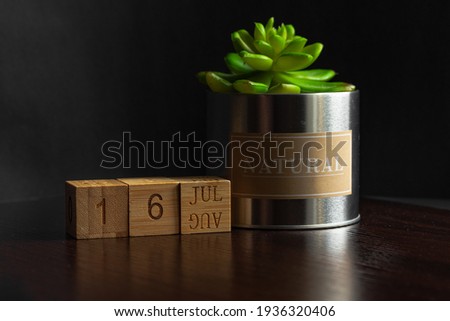 July 16. Image of the calendar July 16 wooden cubes and an artificial plant on a brown wooden table reflection and black background. with empty space for text