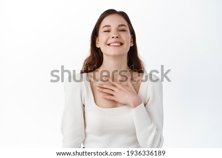 Lovely young brunette female with pleased expression, being touched by heart piercing story, keeps hand on chest, dressed casually, smiles positively, isolated on white background