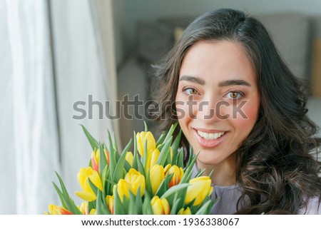 Emotional portrait of young beautiful woman with dark curly hair on light background, holding large bouquet of multicolored tulips in her hands. Spring mood. Mothers Day. Women's Day.